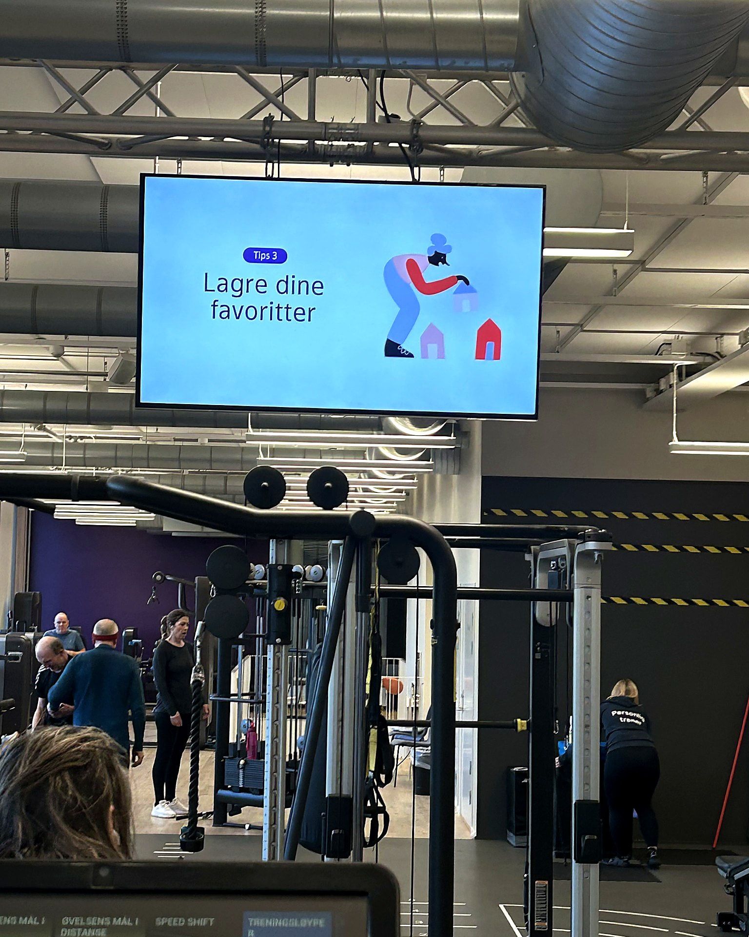 Skjerm i taket over styrketreningsområdet på et treningssenter, med et kort budskap fra senteret.